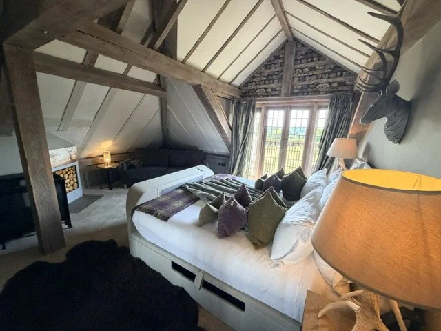 Cozy bedroom with sloped ceiling, wooden beams, a large bed with multiple pillows, a mounted animal head, a side lamp, and a window with curtains showing a scenic view outside.