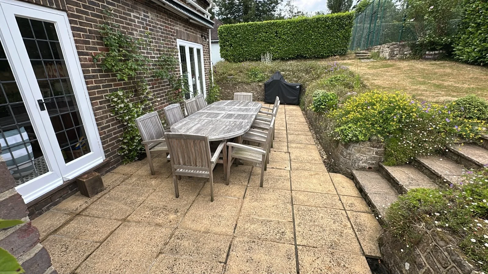 Backyard patio with a wooden table and chairs, a grill covered with a black cover in the corner, a stone staircase, and lush green plants and hedge.