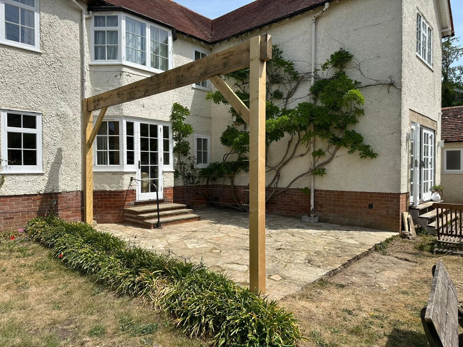 Backyard patio under construction with a wooden frame for a future structure, adjacent to a two-story house with white exterior walls, multiple windows, and steps leading to a glass door. The yard has some greenery and a stone pathway.