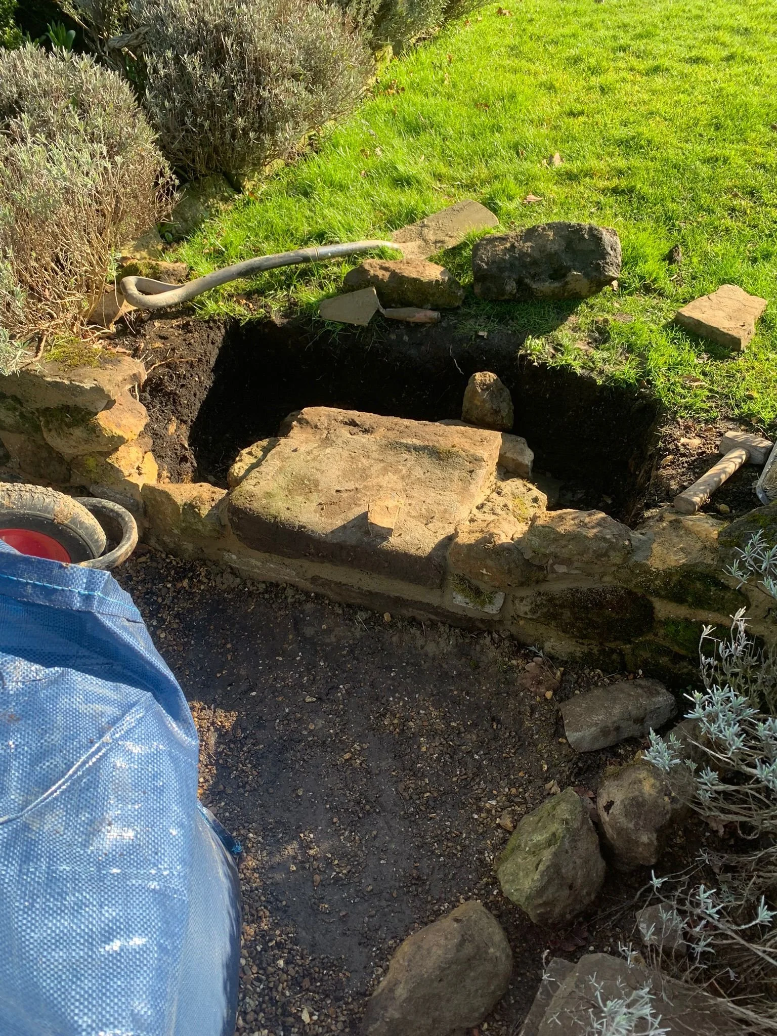 A garden bed is being prepared with rocks placed around the edge and a hole dug in the center. A garden hose is nearby, and green grass surrounds the area. Some plants and bushes are visible to the sides.