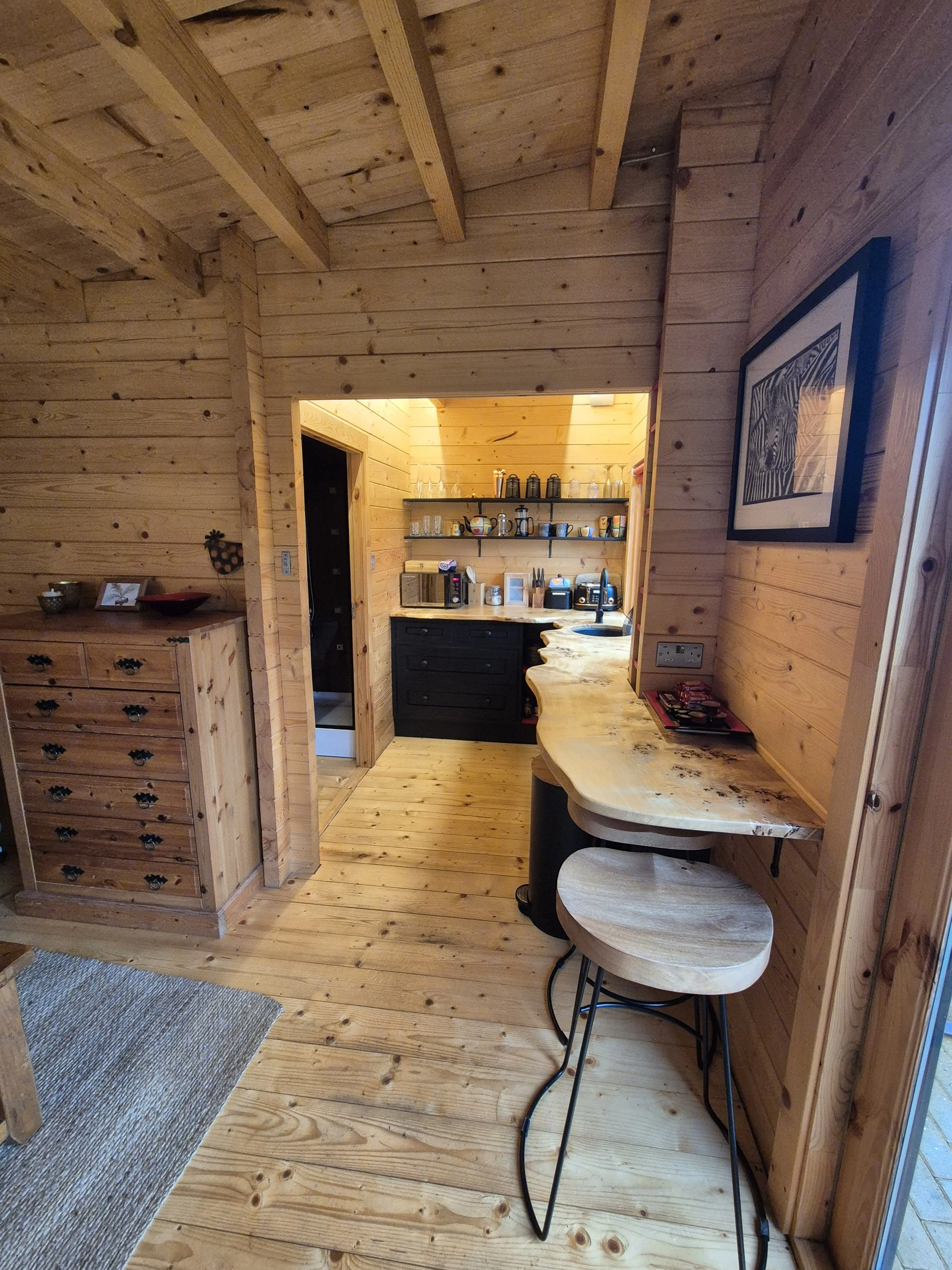 Interior of a wooden cabin kitchen with open shelves, black cabinets, a countertop with appliances, a small wood and metal dining table with two matching chairs, and artwork on the wall.