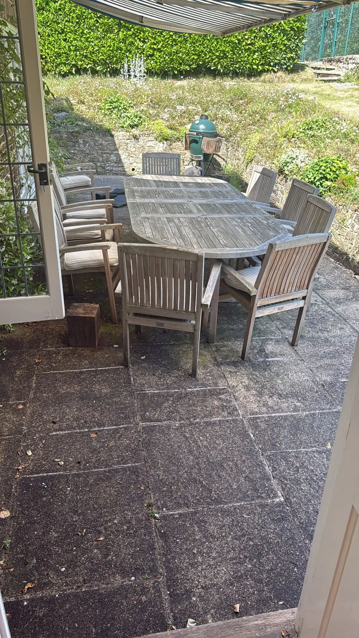 Outdoor patio with a weathered wooden dining table surrounded by multiple wooden chairs, a covered barbecue grill, and a lush green hedge in the background.