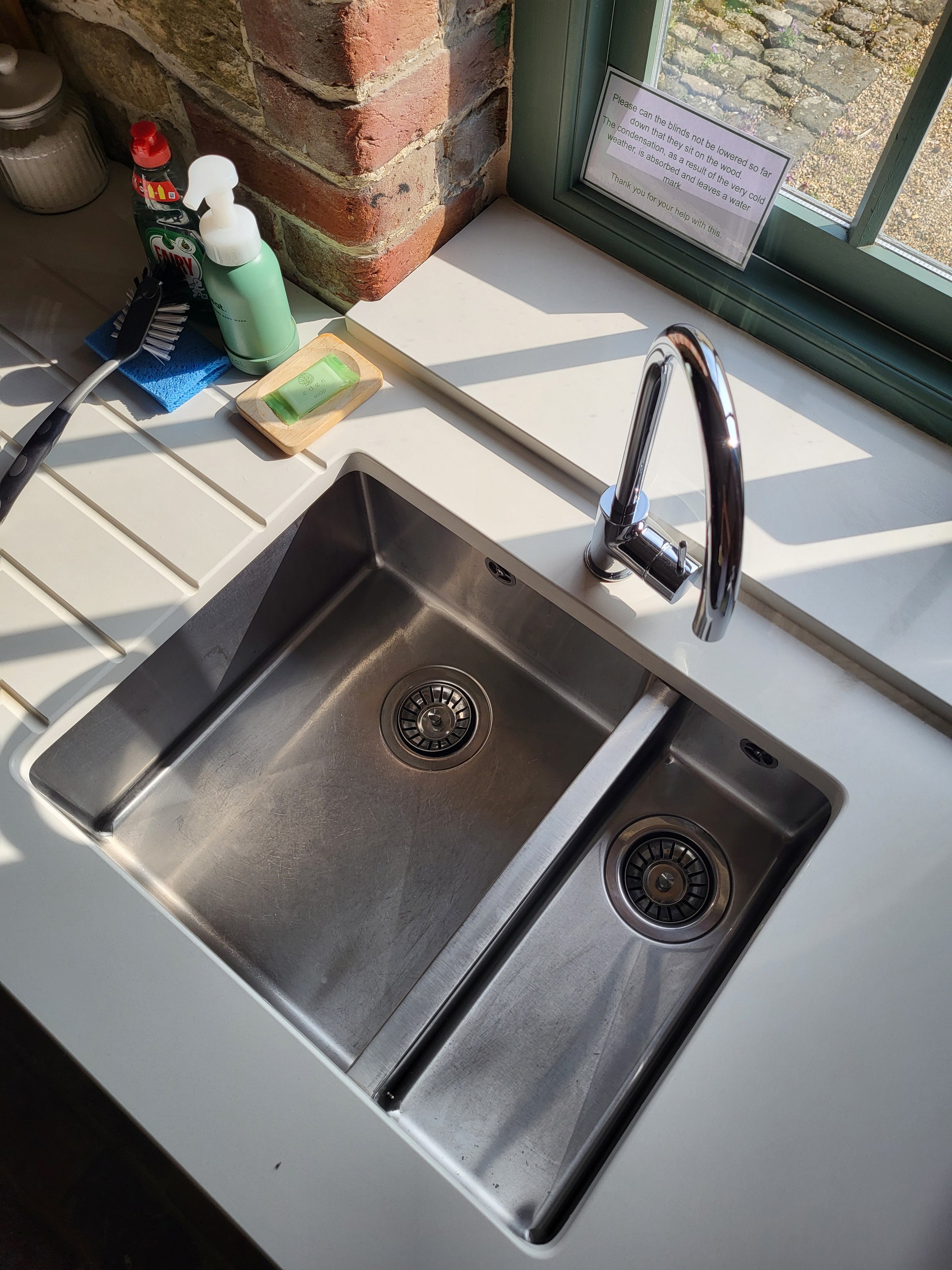 A stainless steel kitchen sink with two basins, a chrome faucet, and surrounding white countertop. To the left of the sink is a soap dispenser, a hairbrush, and cleaning supplies. A window with a sign and a brick wall are visible in the background.