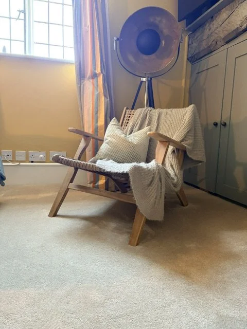 A cozy room corner with a wooden armchair draped with a cream blanket and cushion, a large standing floor lamp, and a colorful curtain by a window.