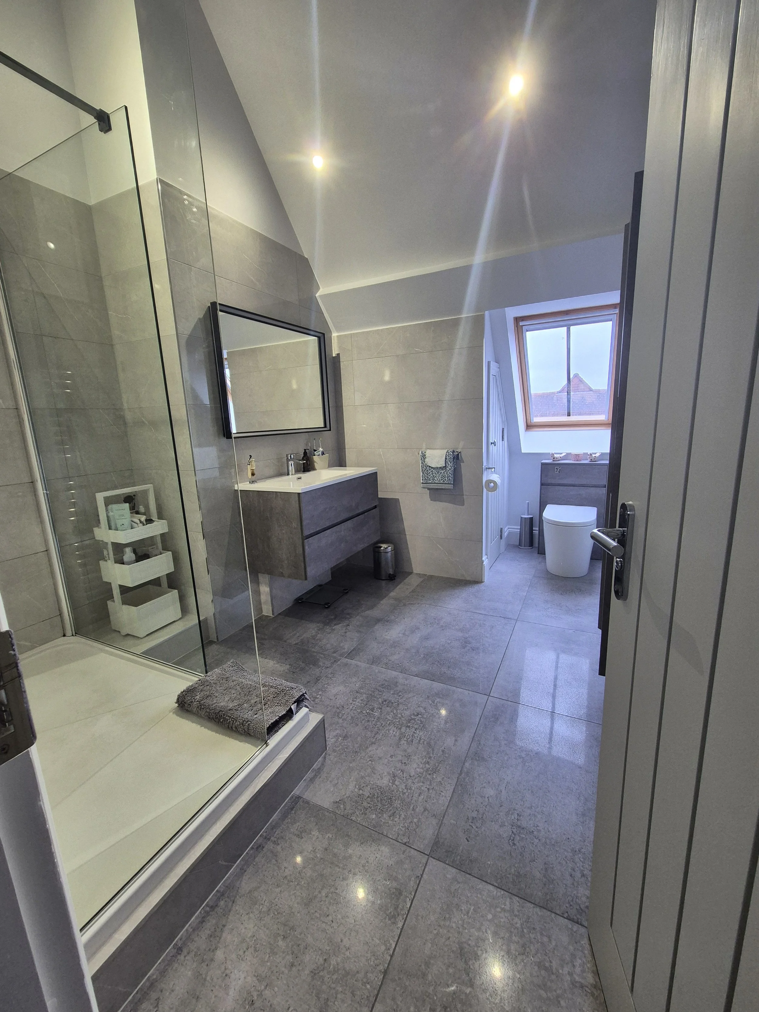 Modern bathroom with gray tiled floor and walls, a glass shower enclosure, a floating vanity with a mirror, and a window with a sloped ceiling.
