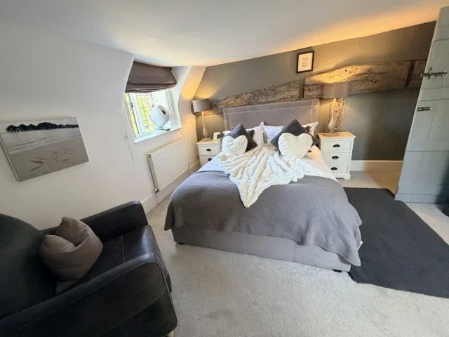 A cozy bedroom with a bed covered in gray bedding, decorated with heart-shaped pillows and a white throw. There are two white nightstands with lamps on either side of the bed, a dark gray wall behind the bed with a rustic wooden accent. To the left, 