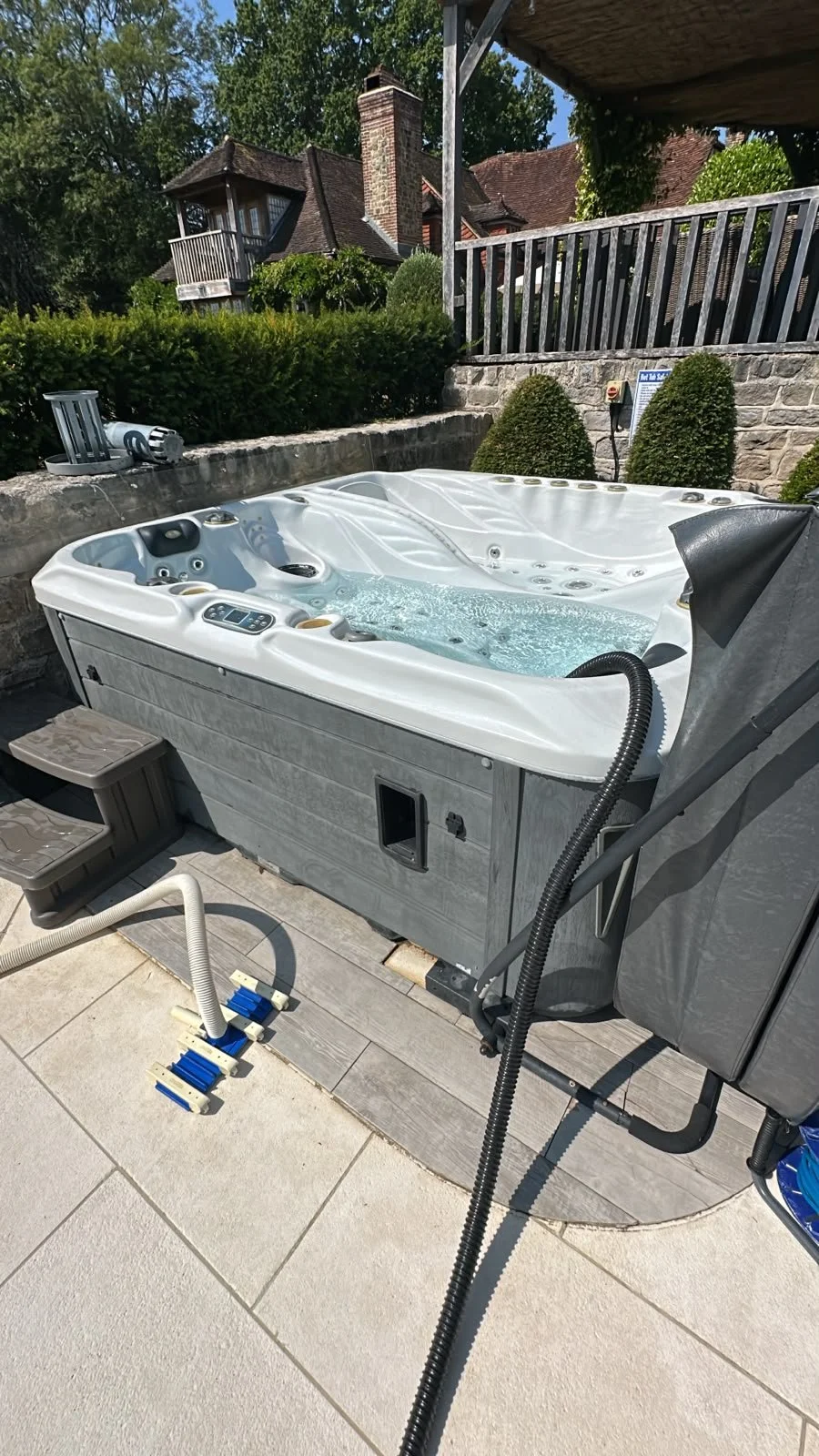 A white outdoor hot tub filled with bubbling water, situated on a patio with stairs and hoses nearby. The background shows a rock wall, bushes, trees, and a house with a brick chimney.