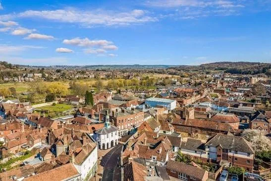 Aerial view of a small town with red-tiled roofs, a church, and surrounding greenery under a blue sky with clouds.