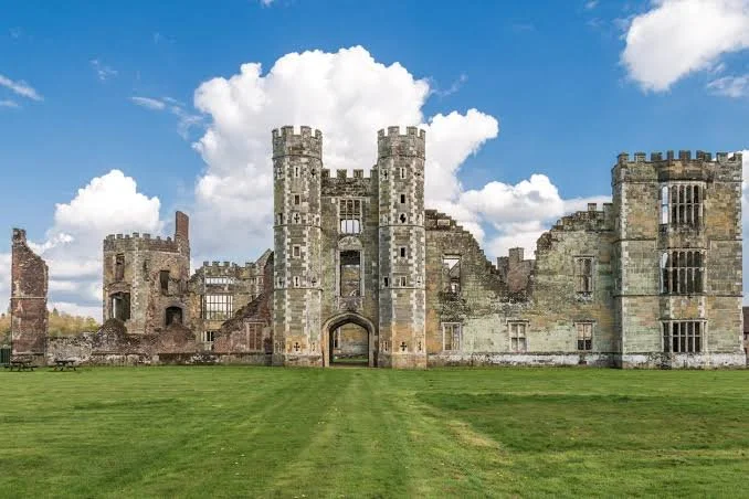 An old stone castle with two central towers, surrounded by green grass and a partly cloudy sky.