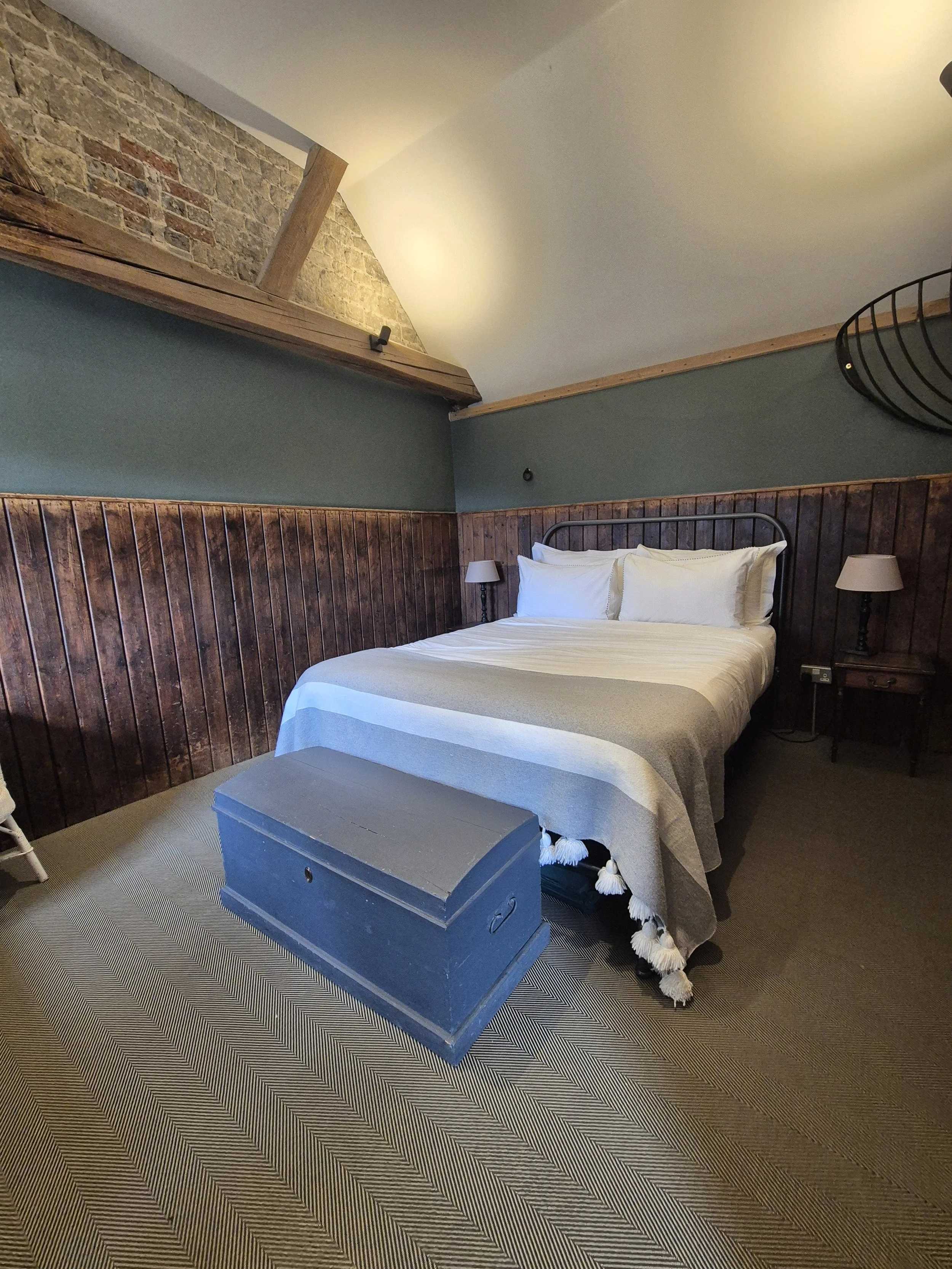 Bedroom with a king-sized bed with white bedding, two matching nightstands with lamps, wooden wall paneling, a stone wall with exposed brick in the upper corner, and a blue storage chest at the foot of the bed. Carpeted floor with a striped pattern.