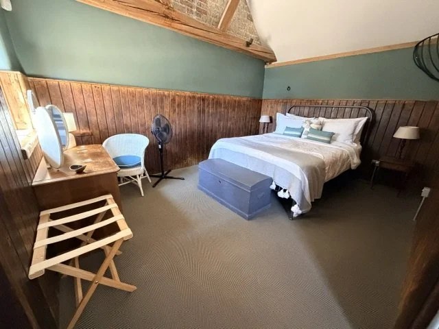 Room with a bed, pillows, nightstands with lamps, a desk with a chair, a fan, and a luggage rack, featuring wood paneling on the lower half of the walls and a sloped ceiling.