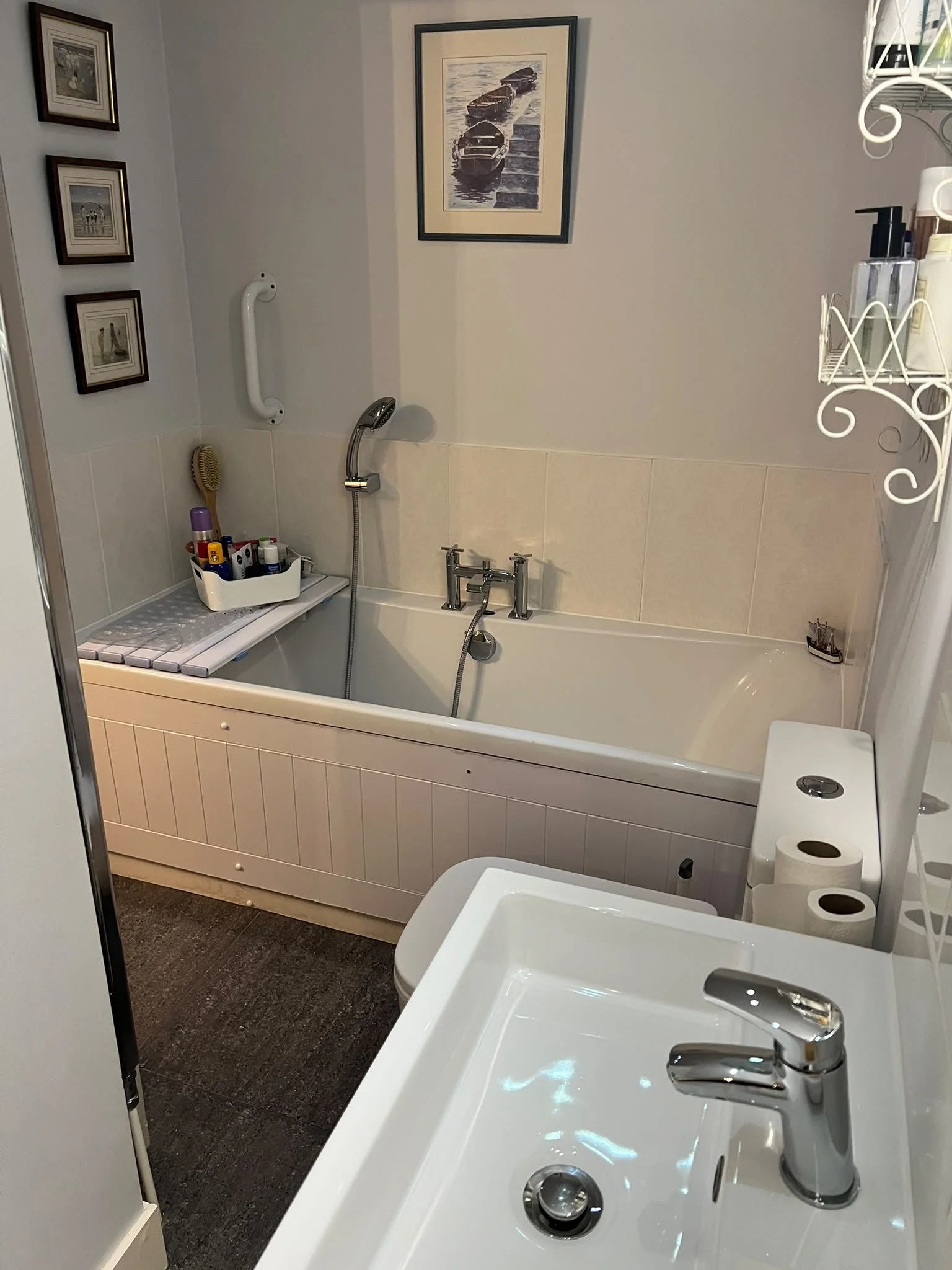 Bathroom with a bathtub, two sinks with silver faucets, a toilet, a small shelf with toiletries, framed pictures on the wall, and a cleaning brush.