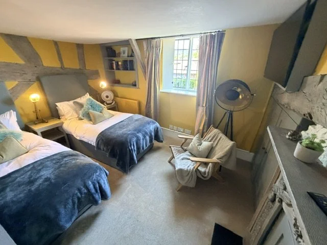A cozy bedroom with two beds covered in blue blankets, each with multiple pillows. There is a window with light-colored curtains, a small armchair with a throw blanket, a vintage fan on a stand, and built-in shelves with books. The room has a warm, i