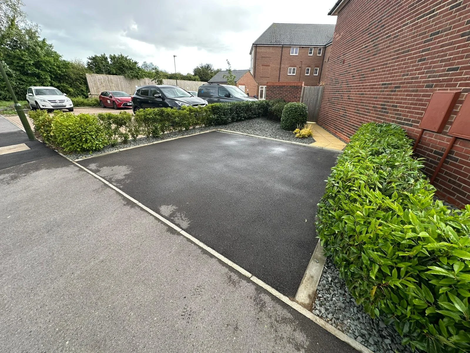 Empty parking space bordered by trimmed green bushes, with other parked cars in the background near a brick building under cloudy sky.