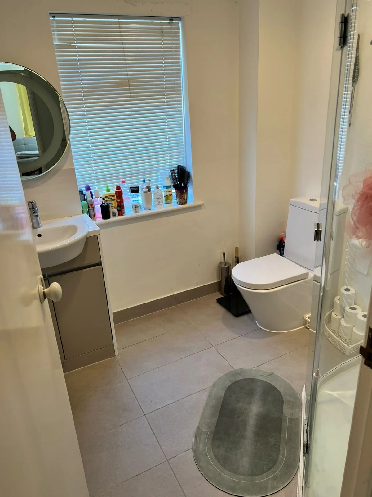 Bathroom with a small sink, a window with blinds, a shelf with toiletries, a toilet, and a shower stall with toilet paper rolls and a gray rug.