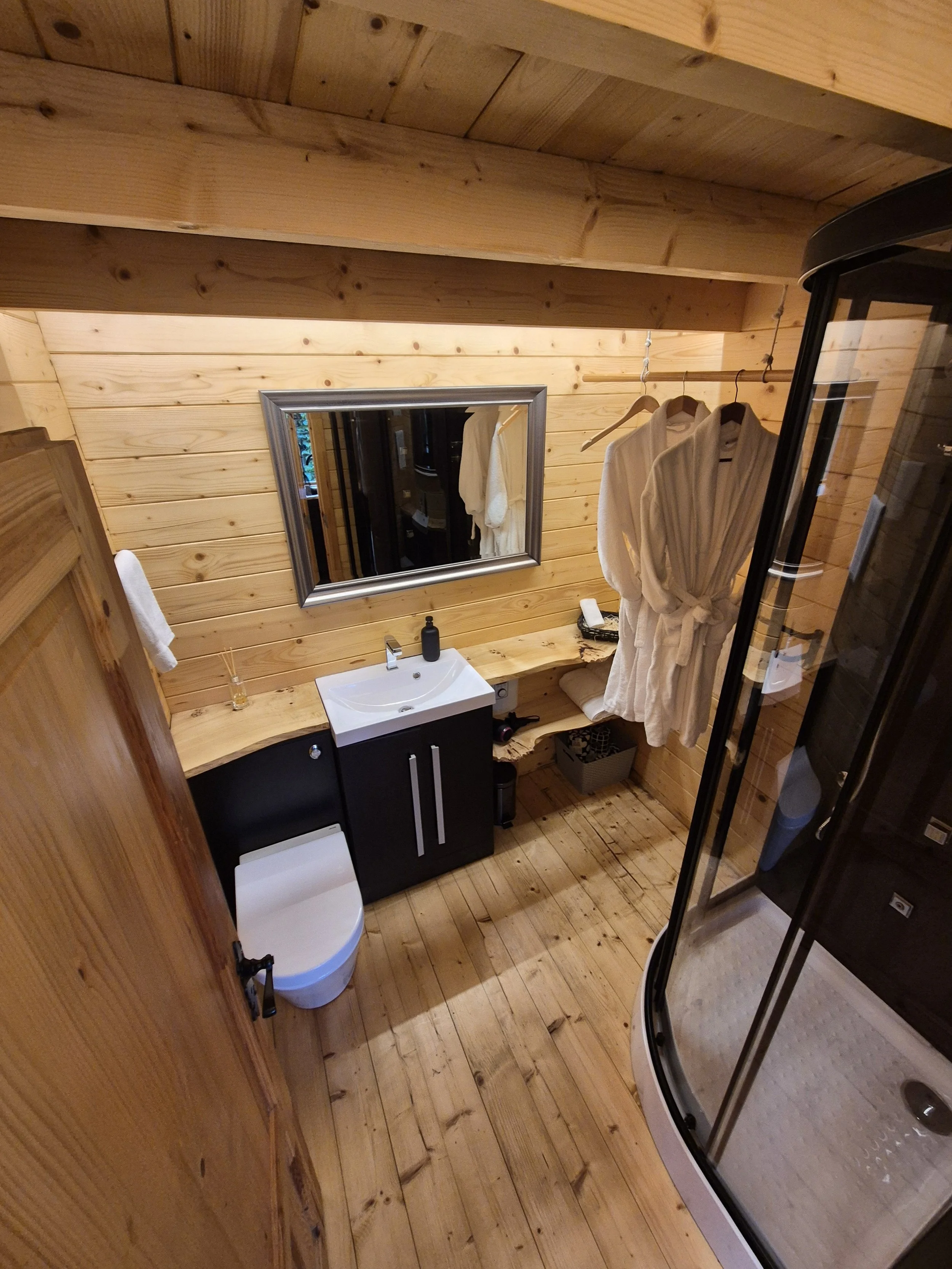 A cozy bathroom with wooden walls and ceiling, featuring a small white sink with a black cabinet, a mirror above it, a white toilet, a glass-enclosed shower, and hanging robes.