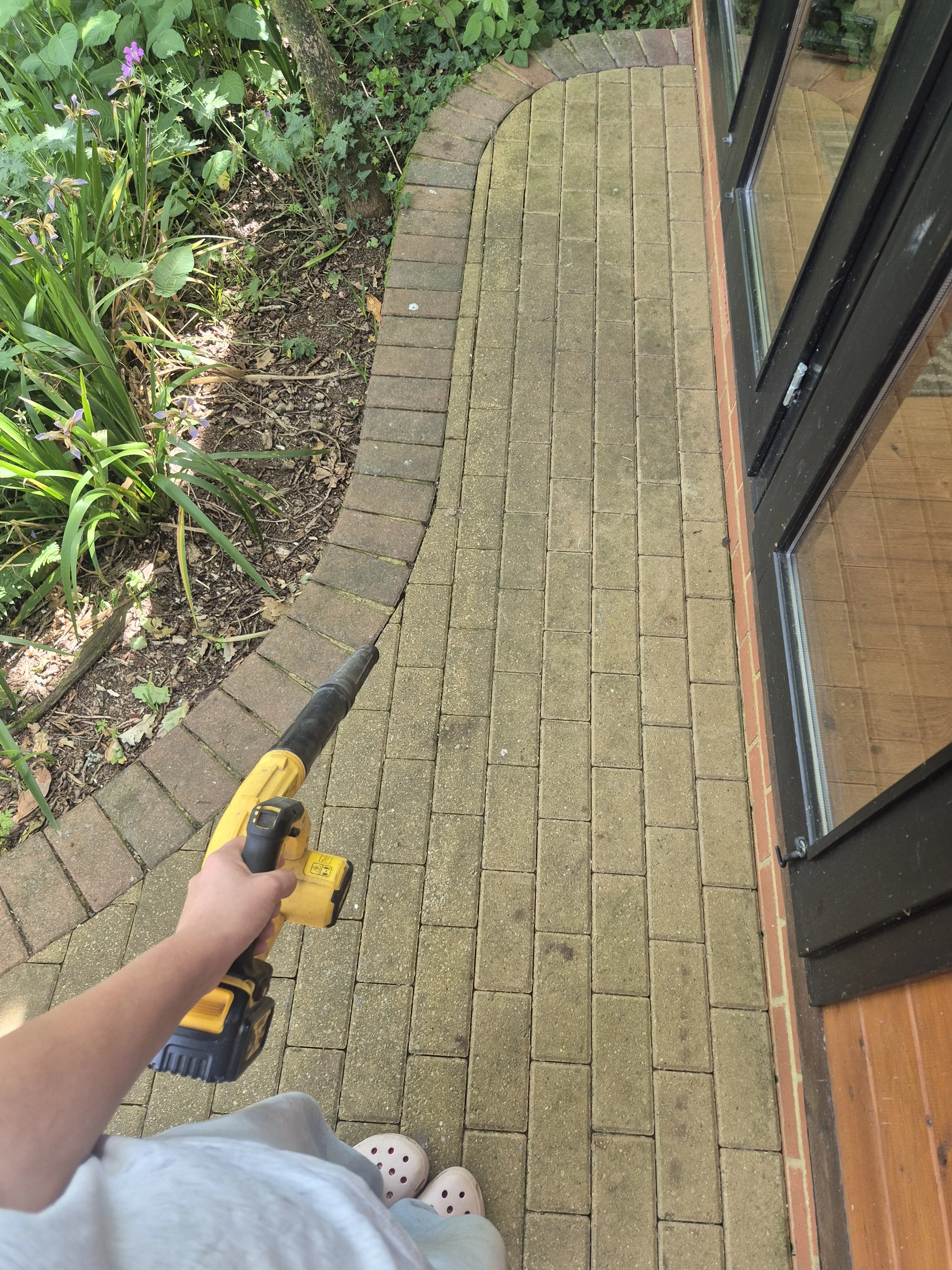 Person holding a yellow cordless drill and walking on a brick patio next to a garden bed with plants and flowers, with a glass door to the right.