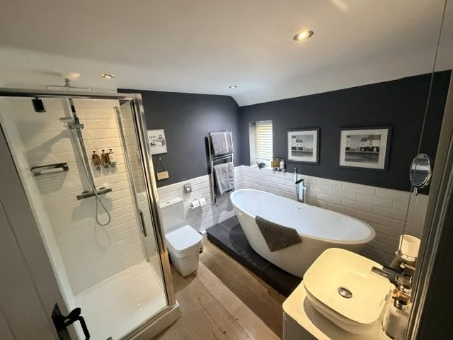 Modern bathroom with a white freestanding bathtub, a glass-enclosed shower, a toilet, and a white vanity sink, with dark accented walls and framed artwork.