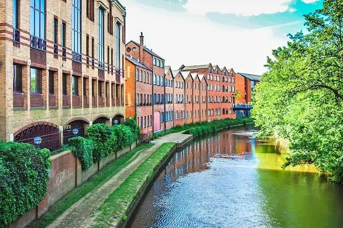 Colorful hotel or apartment buildings along a canal with a brick walkway, lush green bushes, and a large leafy tree under a blue sky.