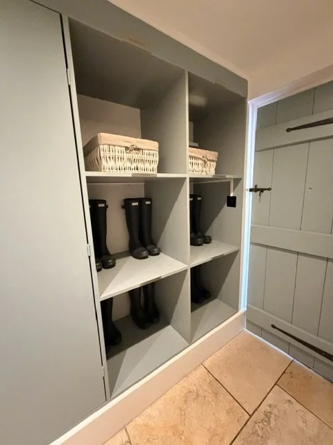 A built-in cubby storage unit with baskets on top and tall black rain boots inside on a shelf, next to a white wooden door with black hinge latch.