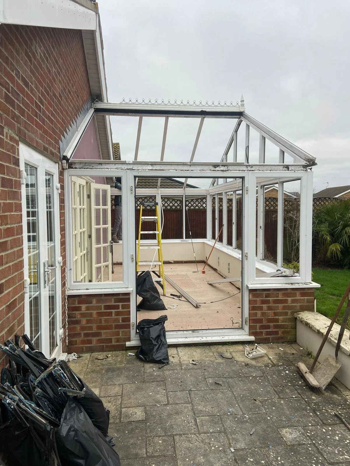 A brick house with an extension under construction, showing a partially built conservatory with a metal frame and glass panels, a yellow ladder inside, and construction tools and debris on the patio.