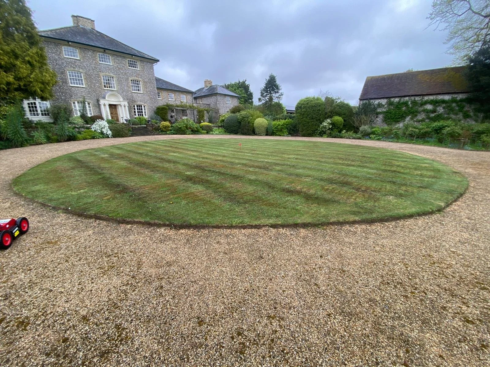 A circular golf green with flagstick in a yard, surrounded by gravel path and a large historic house with stone walls and multiple windows in the background.