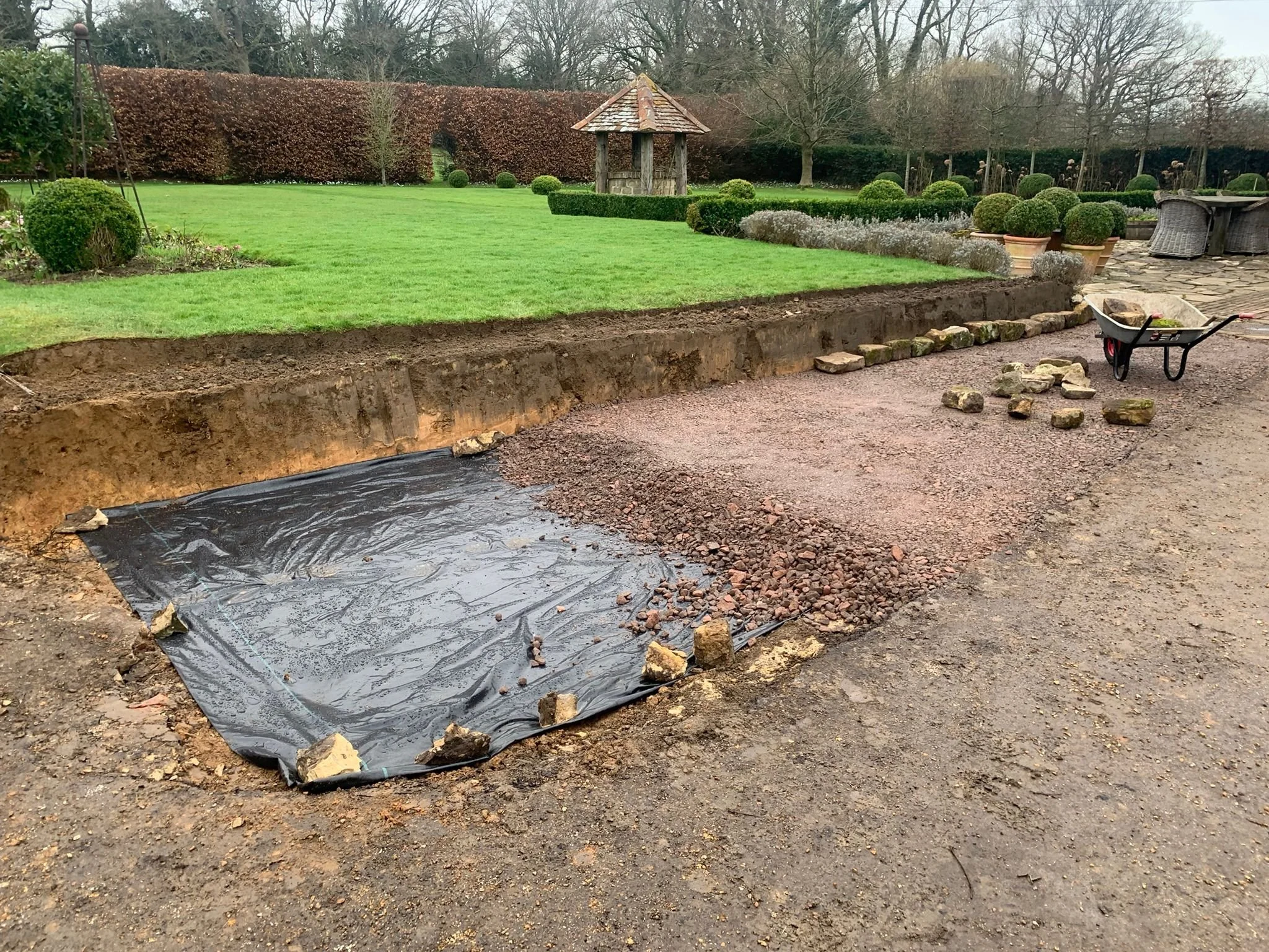 A backyard with a large, lush green lawn, several trimmed bushes, and a stone path. There is a small wooden well with a shingled roof and a wheelbarrow filled with rocks and dirt near the flower bed, as well as some loose stones on the ground. In the