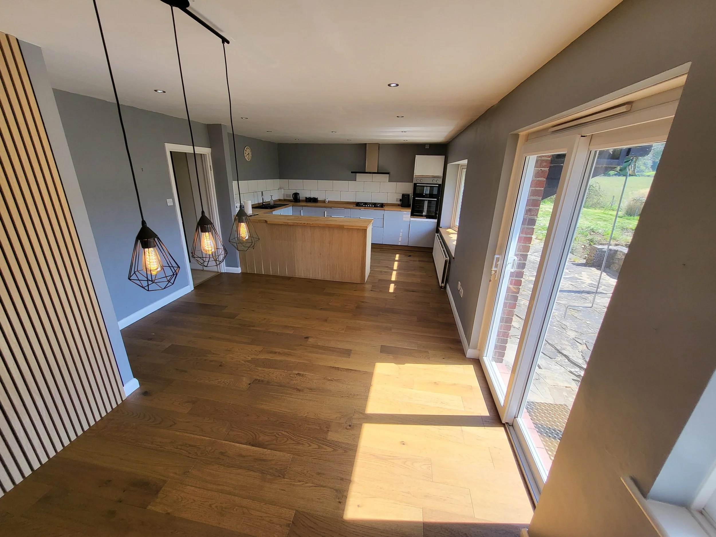 A modern kitchen with gray walls, white tile backsplash, wooden countertops, and light blue cabinets. There are three geometric pendant lights hanging from the ceiling. Large sliding glass doors lead outside to a patio and a yard with a wind chime ha