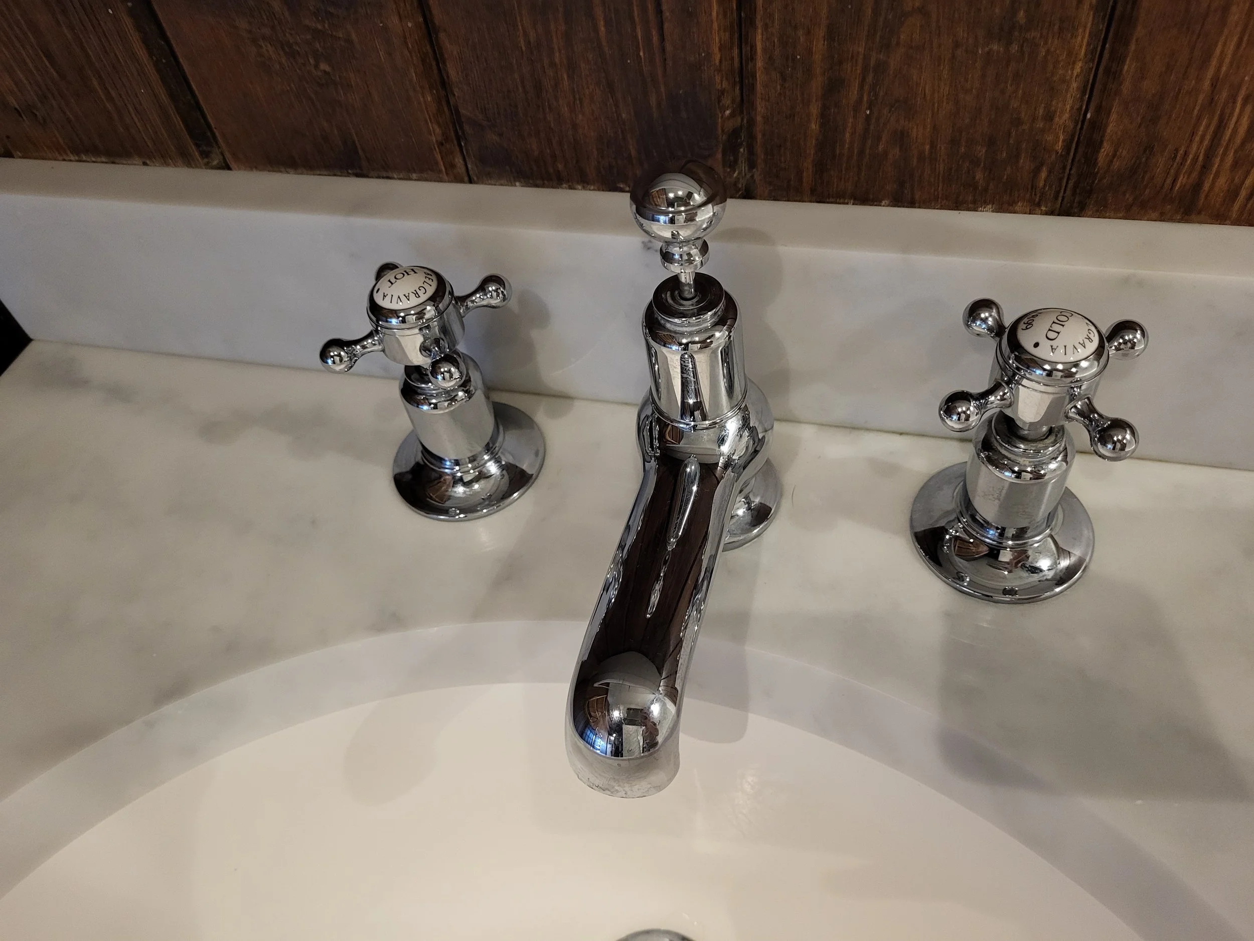 Close-up of a shiny silver faucet with two handles on a white sink, with a wooden wall in the background.