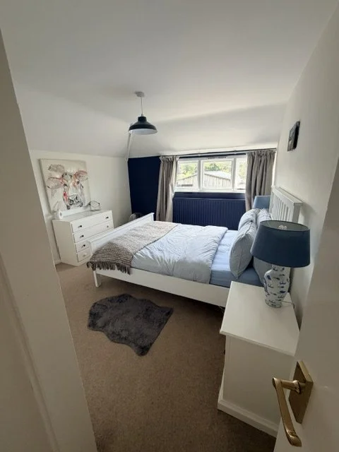 A cozy bedroom with a white bed, blue bedside lamp, white dresser, and a window with curtains overlooking a backyard.