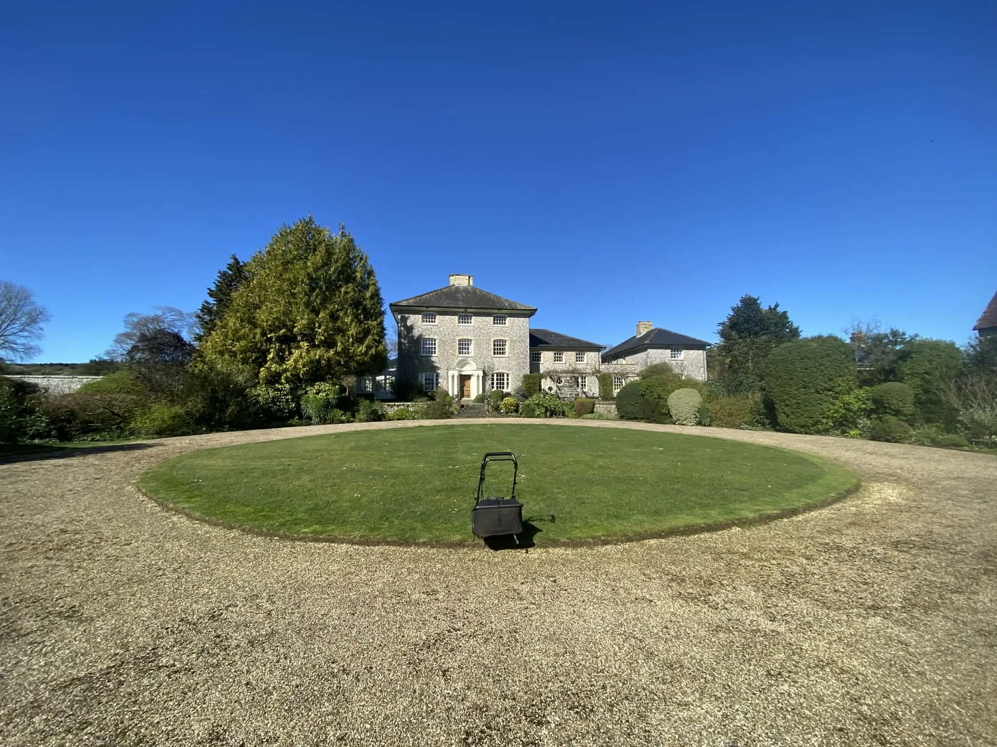 A large house with a stone facade and multiple windows, surrounded by well-maintained trees and shrubbery, under a clear blue sky. A stone pathway or circular driveway is in the foreground with a black rolling suitcase on it.