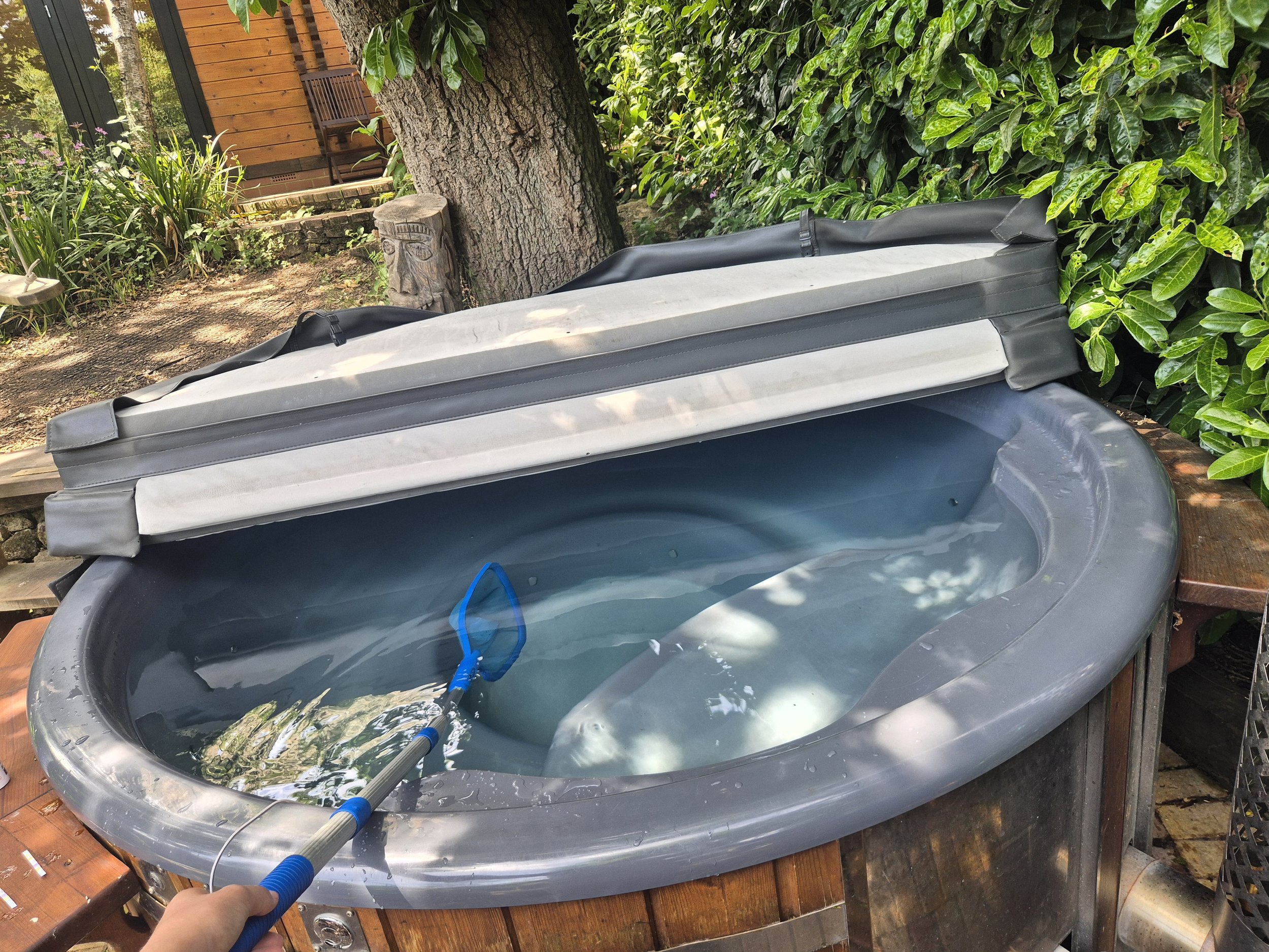A hot tub filled with water outdoors next to a tree and green hedges, with a wooden house in the background. A person's hand is visible holding a pool cleaning tool.