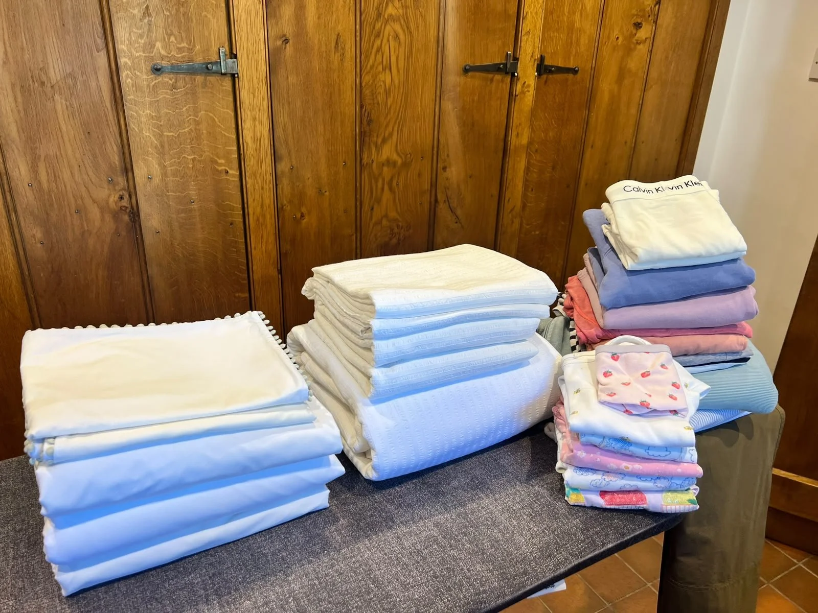 Stacks of folded white, pastel, and patterned diapers or cloths are arranged on a table against a wooden wall.