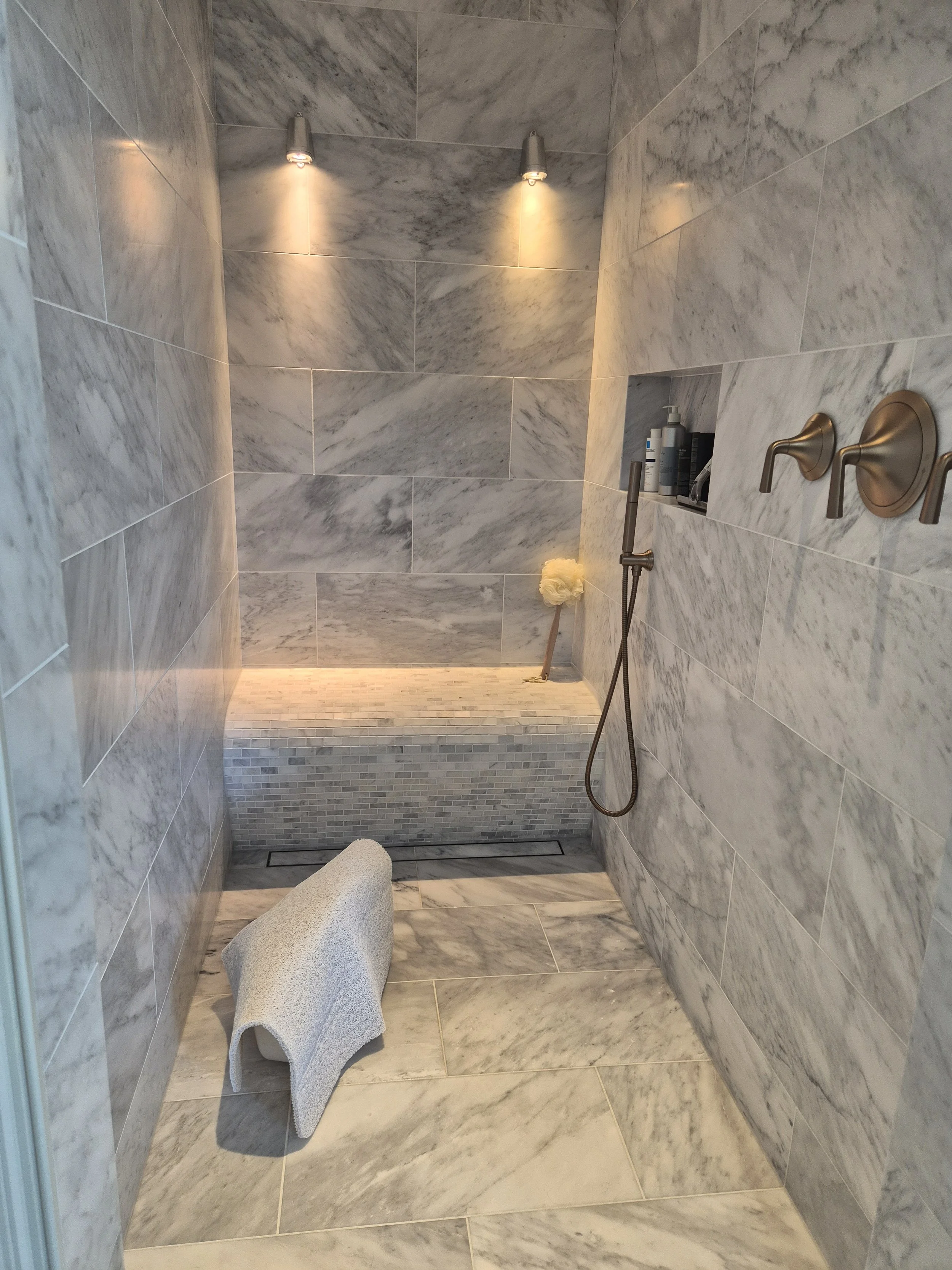 A modern shower room with marble tiles on the walls and floor, featuring two wall-mounted shower heads, a built-in shelf with toiletries, a handheld shower, and a rolled towel on a textured stool.