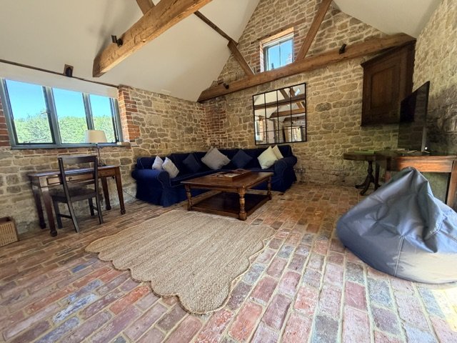 Living room with brick floor and walls, wooden beams, large window, blue sofa with white pillows, wooden coffee table, desk with chair, TV on a stand, and a gray bean bag chair.
