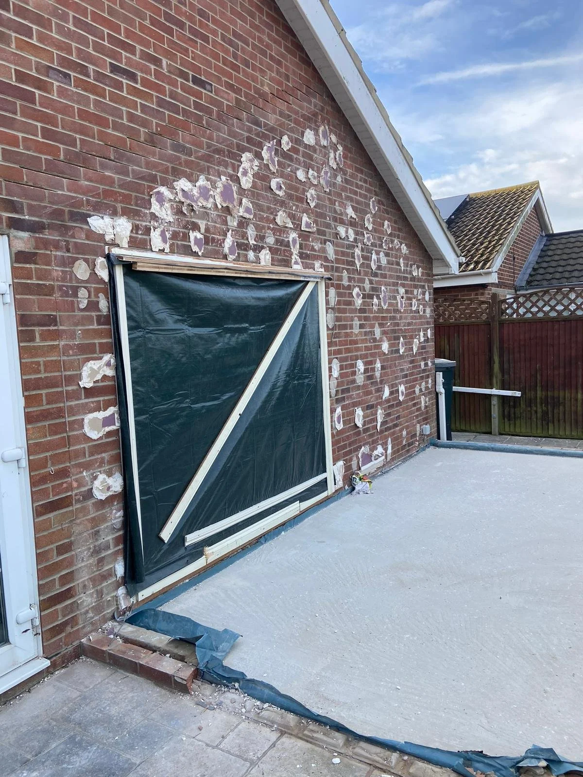A brick house wall with paint peeling and a plastic-covered window frame propped against it. There's a concrete patio and a wooden fence in the background.