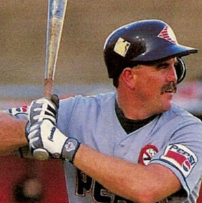 A baseball player at bat, wearing a helmet, sunglasses, and a grey jersey with Pepsi logos, holding a bat in a ready stance.