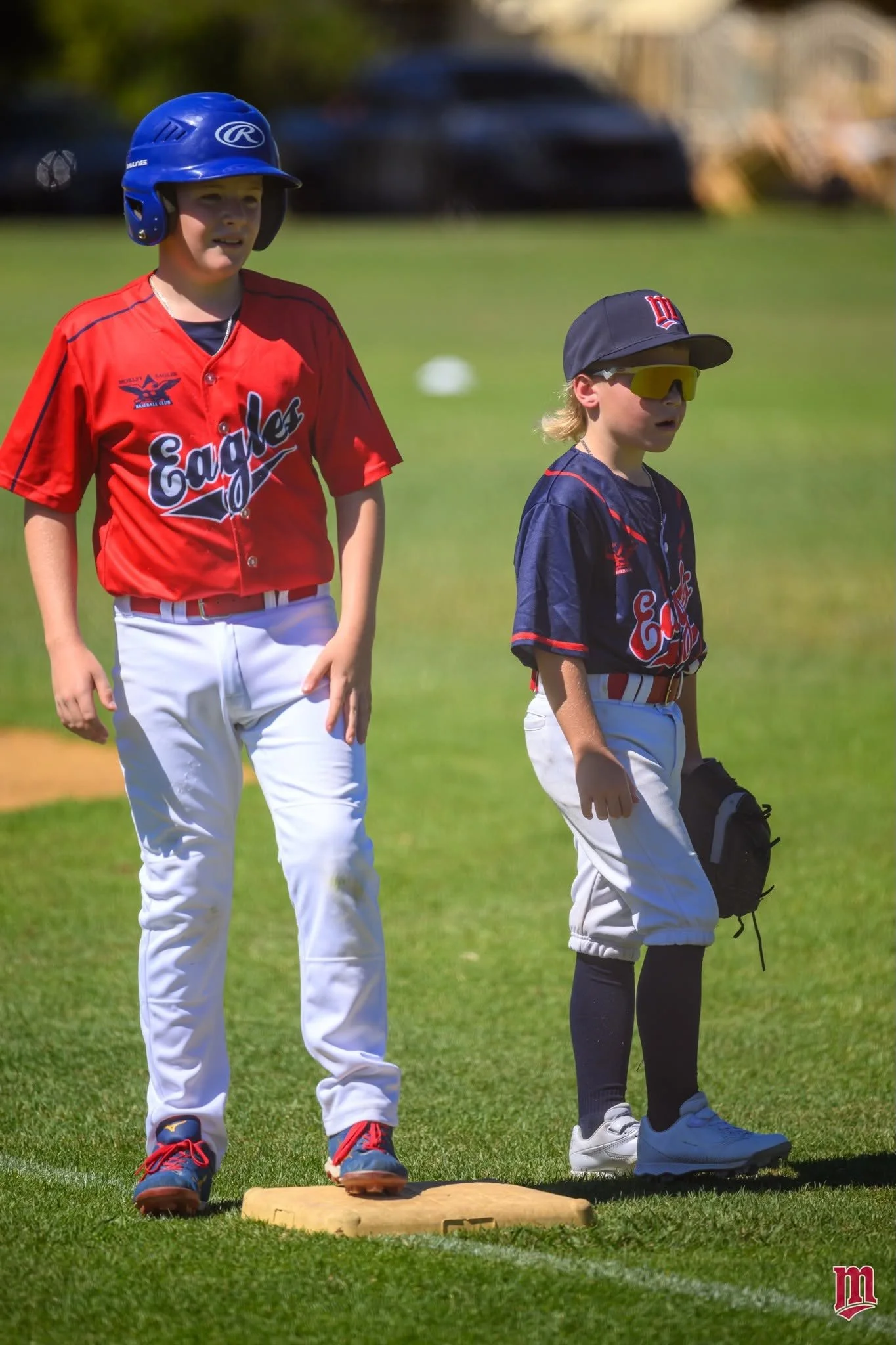 Two young baseball players standing on a field. The player on the left is wearing a red jersey, white pants, a blue helmet, and red and blue shoes. The player on the right is wearing a black jersey, white pants, a black cap, yellow sunglasses, and black knee-high socks, holding a black glove. They are standing near a base on the grass in daylight.