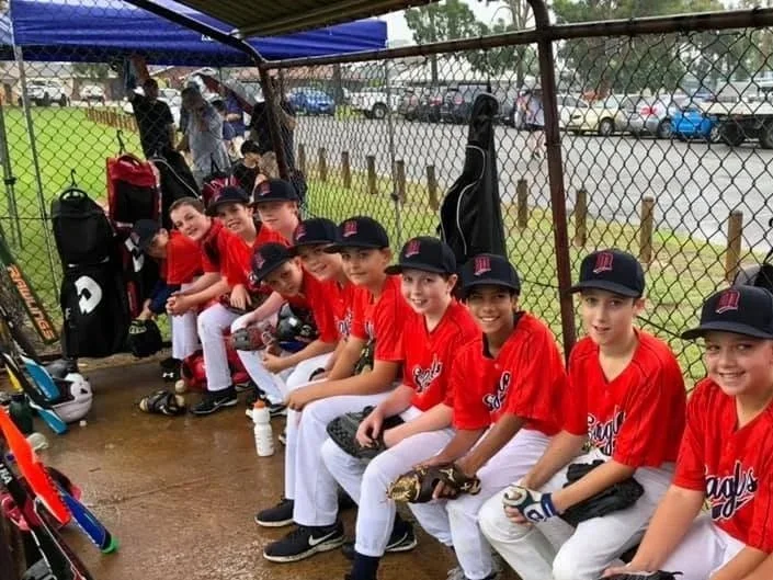 Young female and male baseball players in red jerseys and black caps sitting on a bench in a baseball dugout, smiling and holding baseball gloves and equipment.