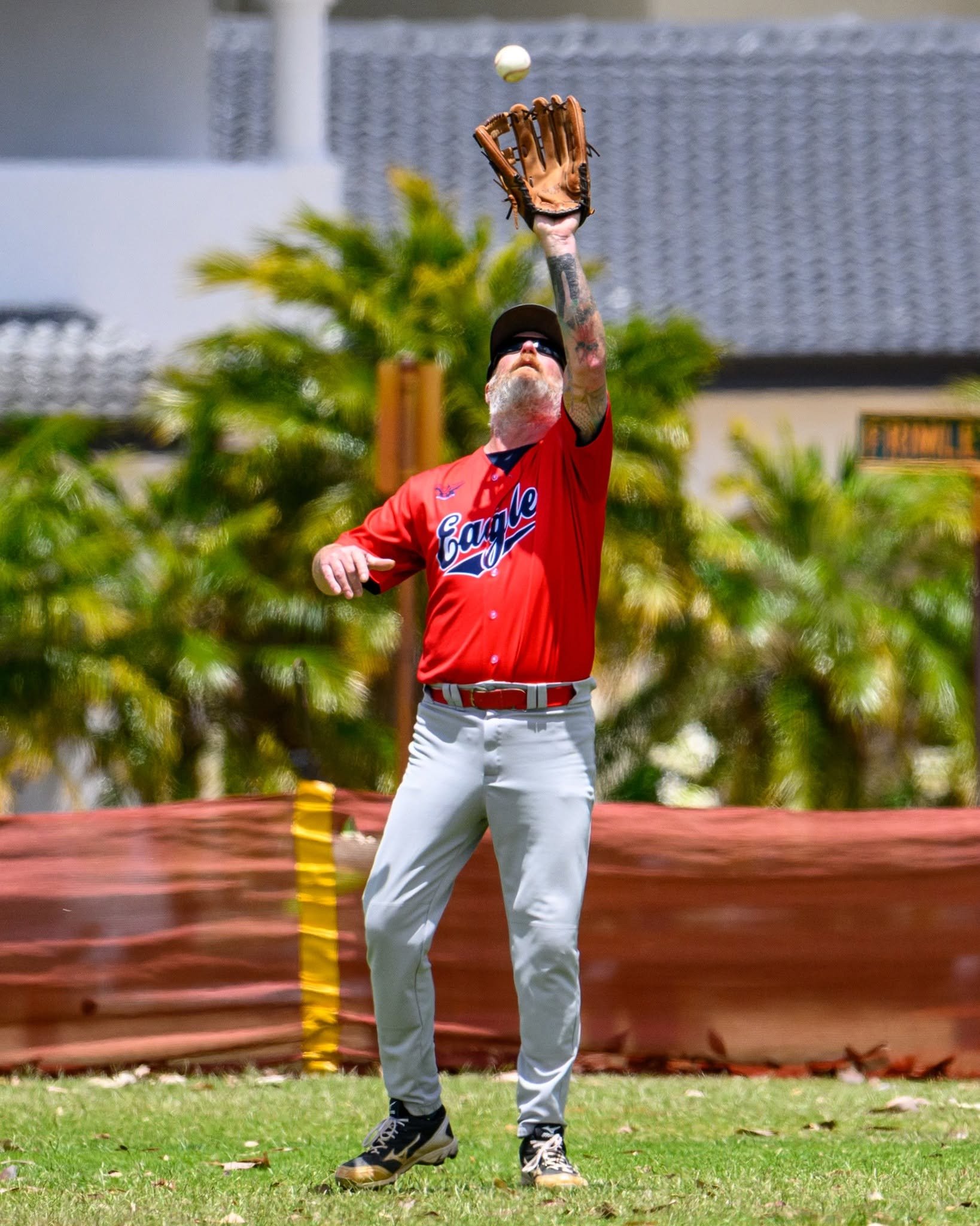 A man wearing a red baseball jersey, white pants, and sneakers is playing baseball, reaching upward to catch a ball with a glove.