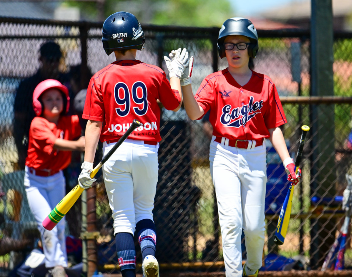 Two young baseball players in red jerseys giving a high five on the field, with a girl in a helmet and red uniform in the background near a chain-link fence.
