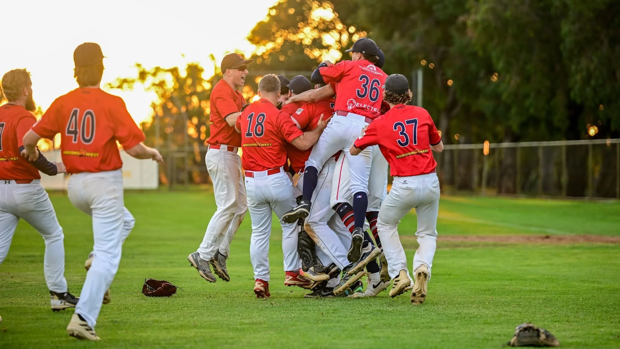 Baseball players in red jerseys celebrate on the field during sunset, with some jumping and hugging.