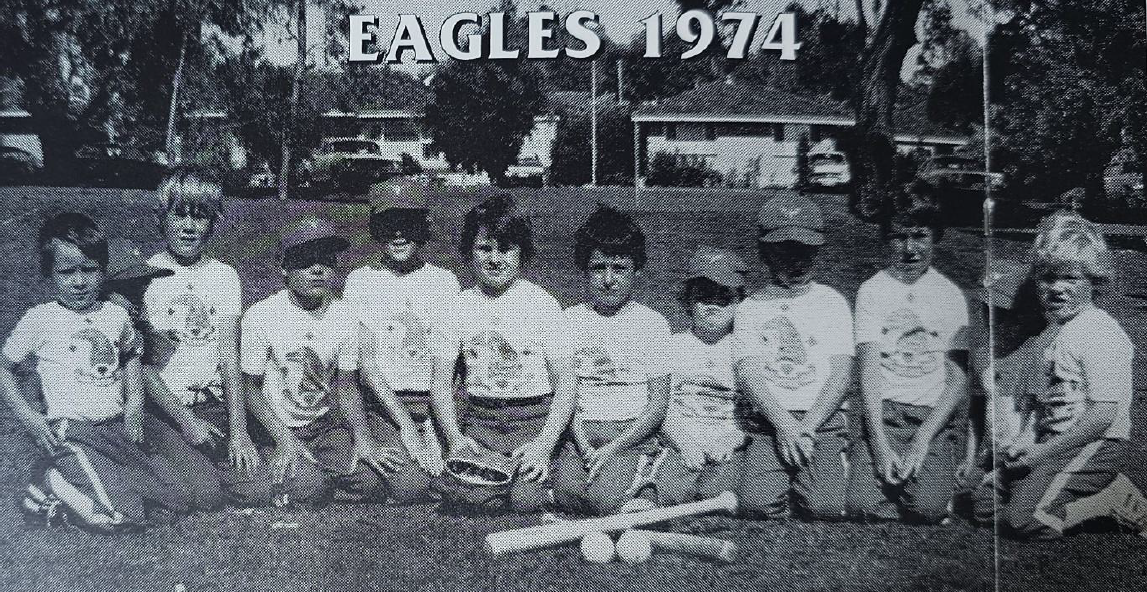 A black and white photo of a youth baseball team named 'Eagles 1974' with ten kids kneeling on the field, wearing baseball uniforms, baseball caps, and smiling, with a baseball bat and ball in front of them.