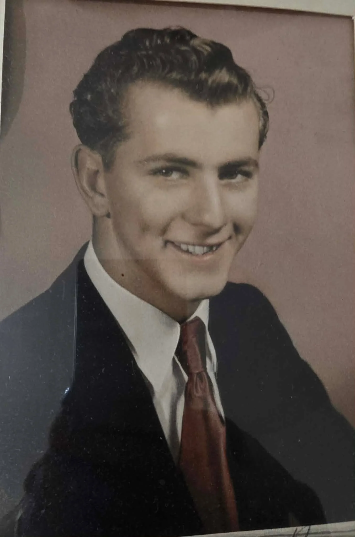 A black and white photo of a young man with wavy hair, dressed in a tuxedo with a white shirt and a patterned tie, smiling.