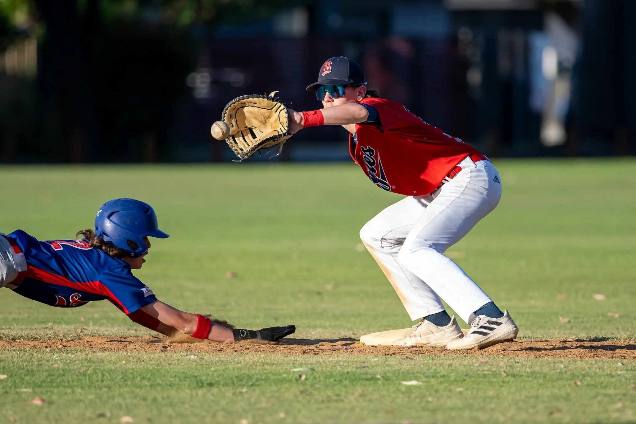 A baseball player in a red jersey and white pants reaches out to catch a sliding base runner during a game, both players wearing caps and sunglasses, on a grassy field.