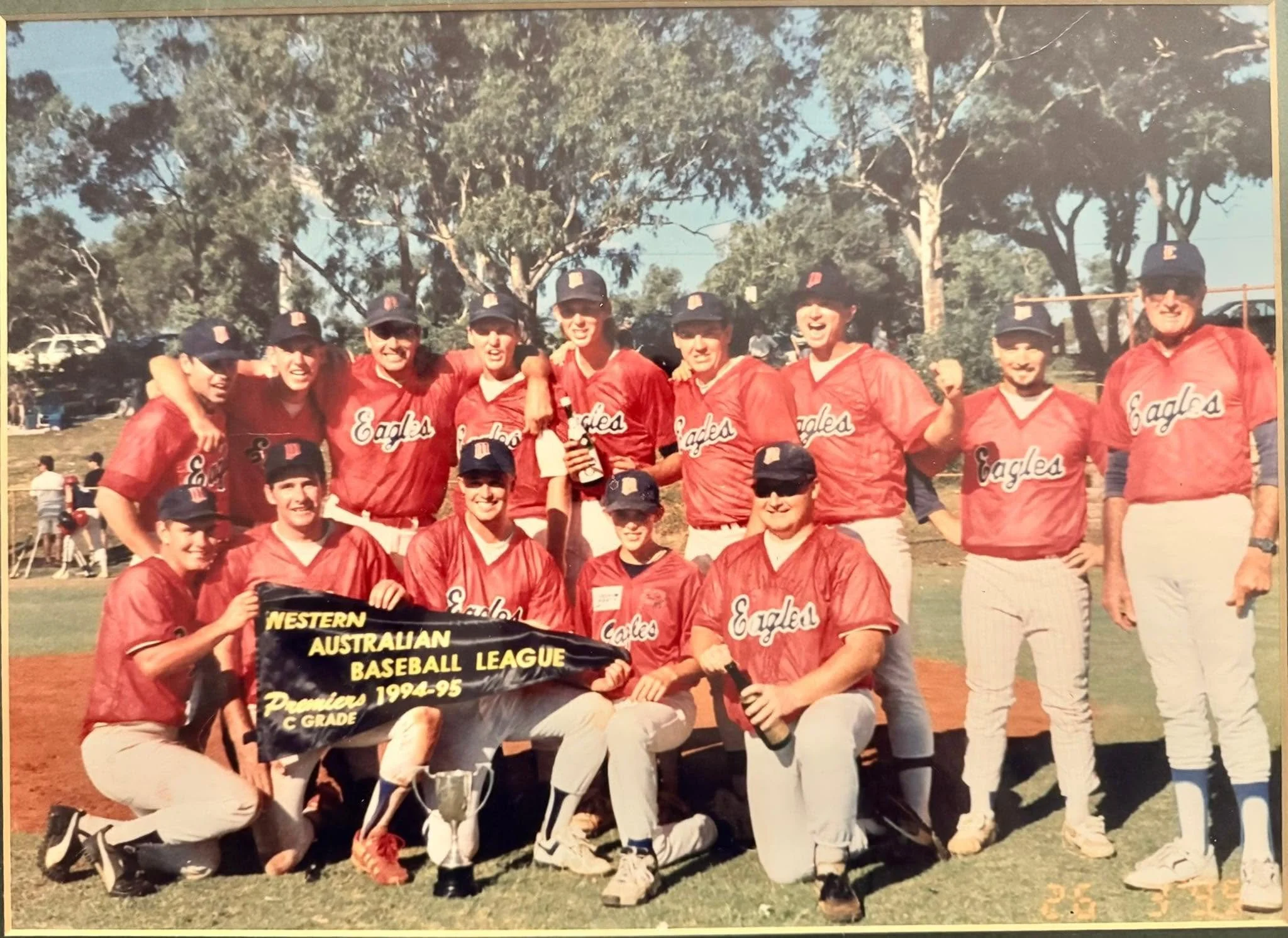 A group of young baseball players in red uniforms and caps, standing outdoors on a baseball field, holding a banner that reads 'Western Australian Baseball League Premier 1994-95'.