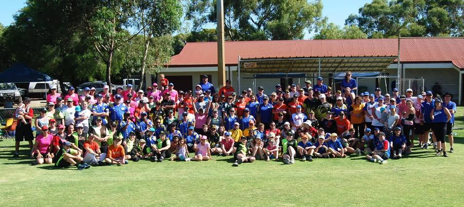 Large group of children and adults gathered outdoors on a baseball field for a group photo, with trees and a building in the background.