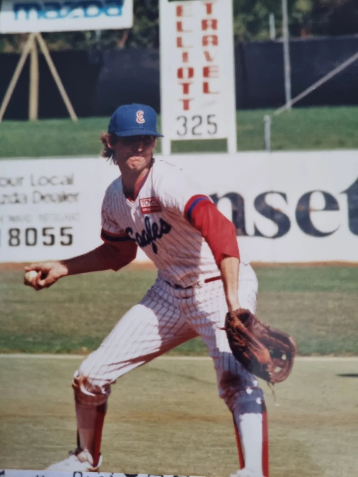 A baseball player in a pinstriped uniform and blue cap with a red and white logo, fielding a ball on the grass outfield with a glove, during a game at a baseball stadium.