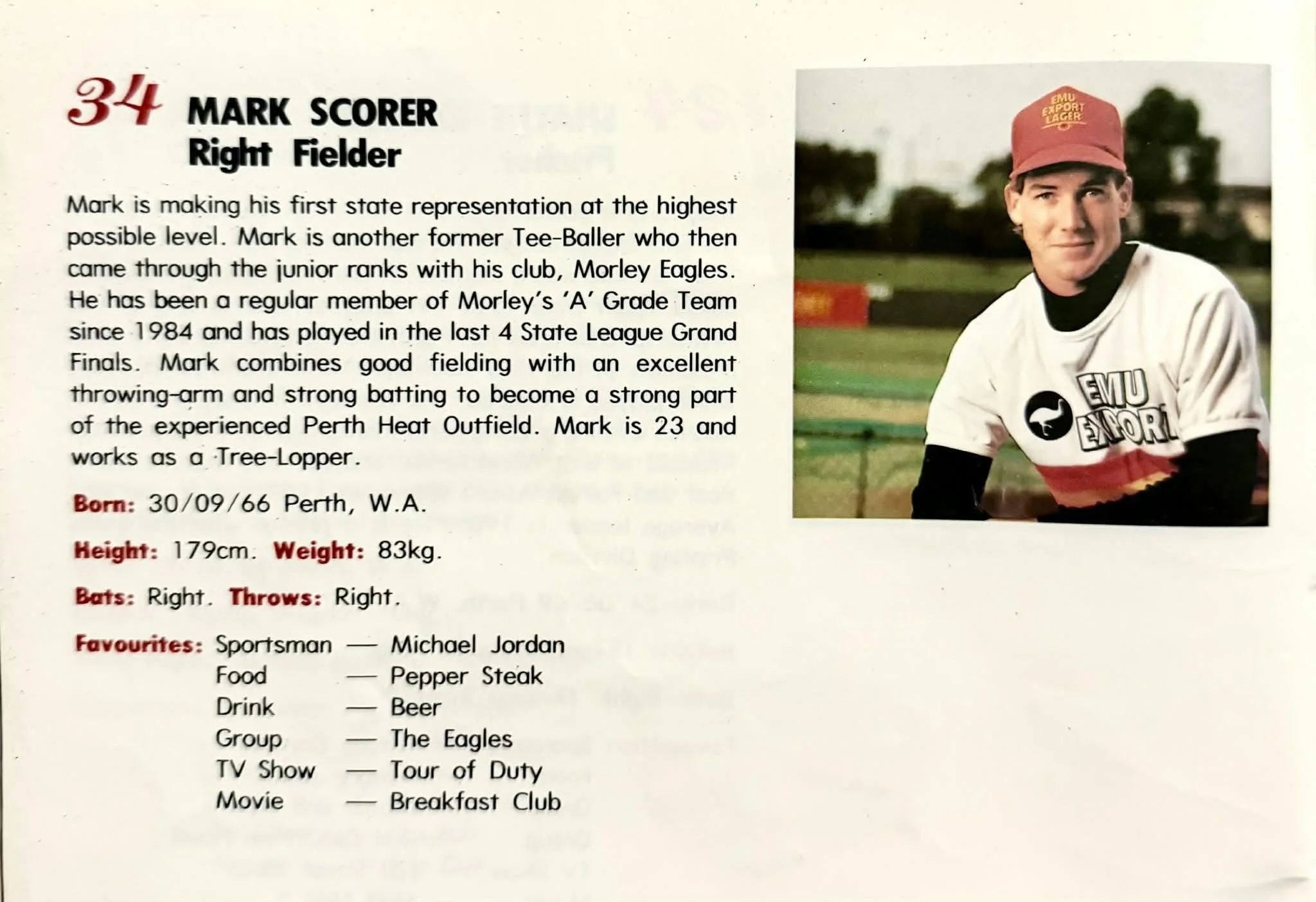 Photograph of a young male athlete in a sports uniform, wearing a red cap with yellow print and a white jersey with black, red, and yellow accents, sitting outdoors on a sports field with trees and a cloudy sky in the background.