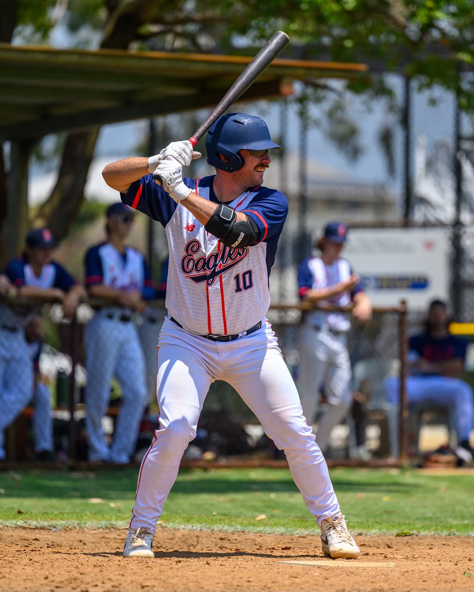 A baseball player at bat, wearing a navy blue helmet, sunglasses, white uniform with red and blue accents, and a wristband, standing on a dirt basepath. In the background, spectators sit behind a fence, with green trees and a blue sky nearby.